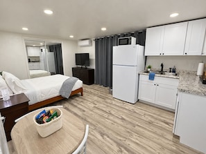 A compact hotel room with a bed, bedside table, TV, kitchenette, and a small dining area.