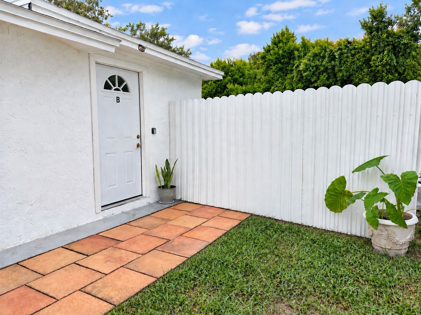 A white house with a door marked 'B', a white picket fence, and a small garden area with grass and a potted plant.