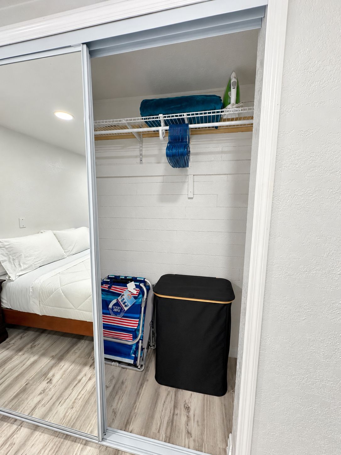 A room with a bed, a suitcase, a small black ottoman, and a blue suitcase with red and white stripes.
