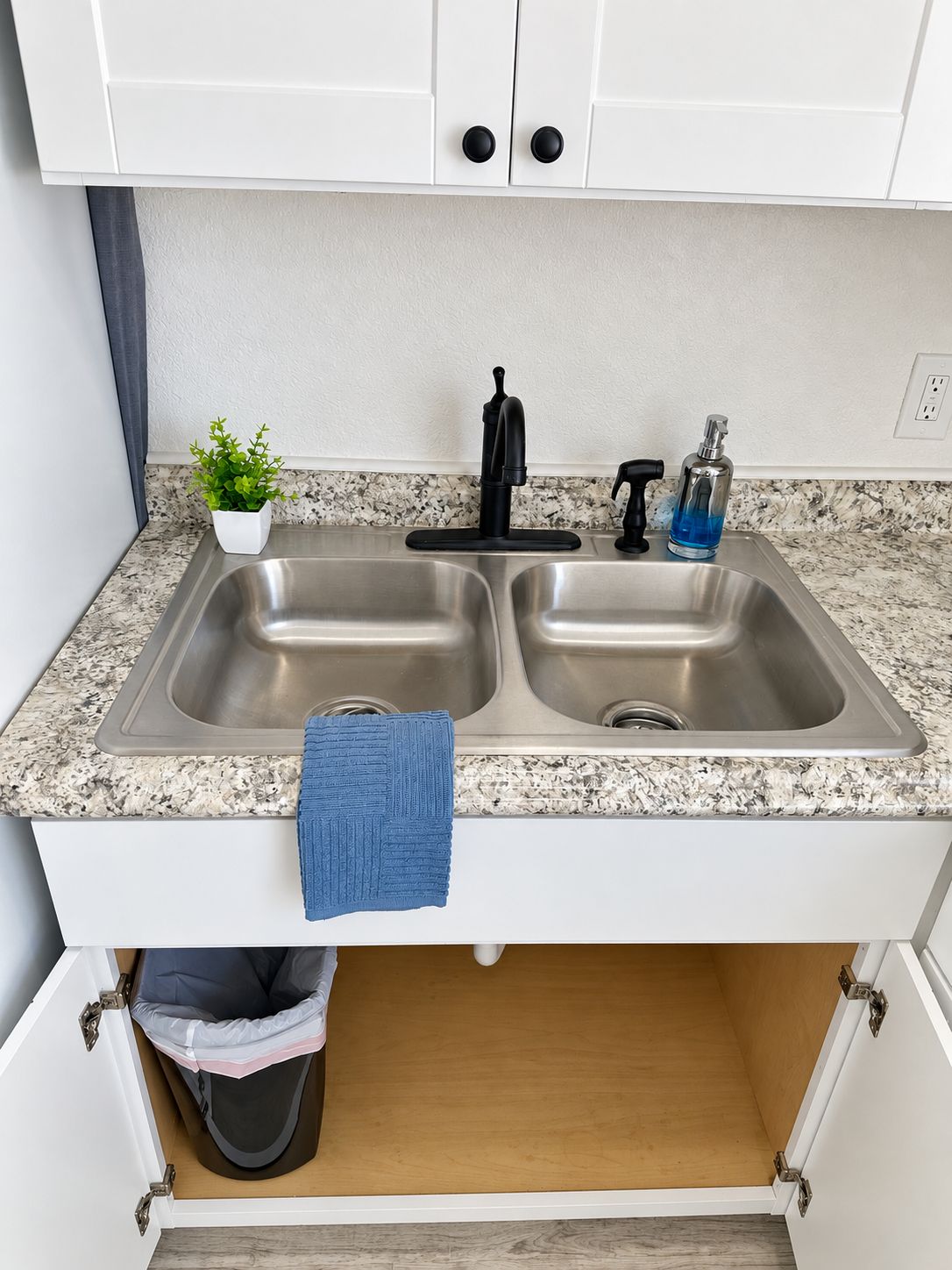 A kitchen with a double sink, a potted plant, a blue towel, and a trash bin.