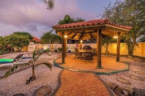 A covered outdoor seating area with a tiled roof, surrounded by a gravel path and small plants. - SunnyStay Villa - A Spacious 5-Bedroom Villa with Pool & Mini Golf (Moko)