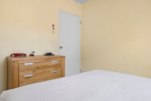 Een slaapkamer met een houten dressoir, een bed met een geweven witte spreideken, en een witte deur met een slot en een rood bord met daarop een nooduitgang.