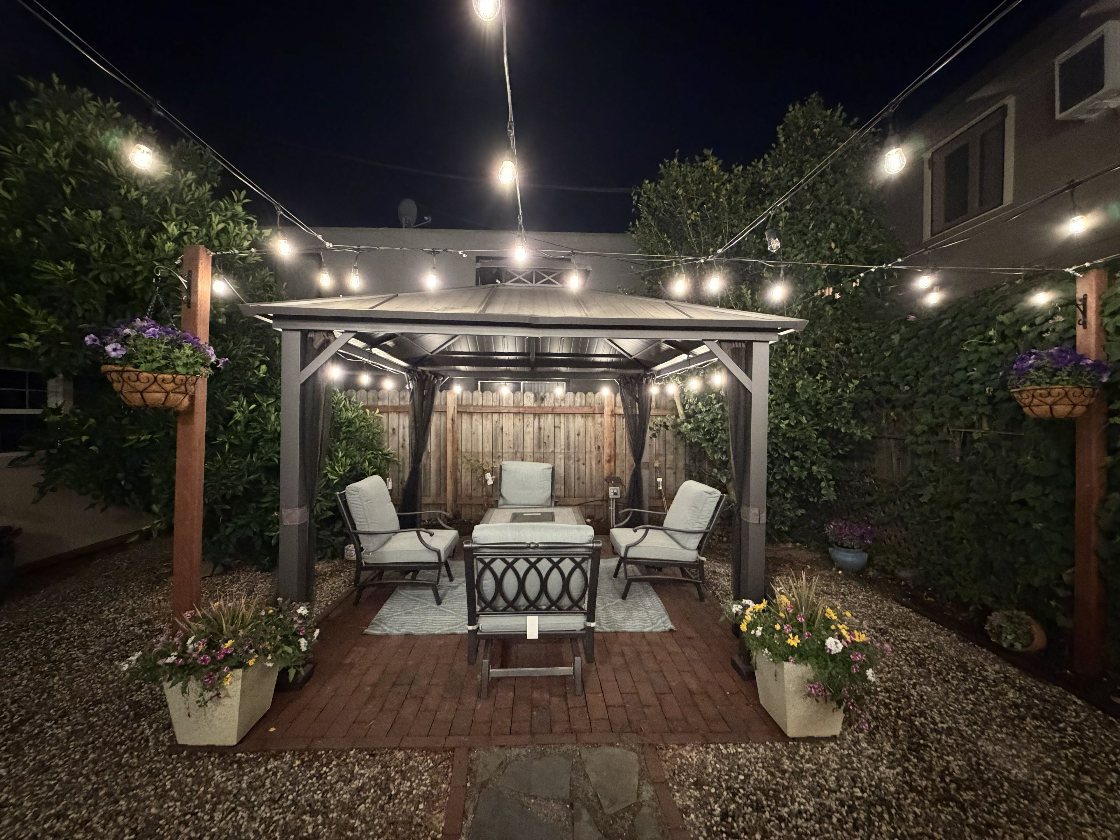 Une terrasse couverte avec une table et des chaises, entourée de plantes en pot et illuminée de guirlandes lumineuses.