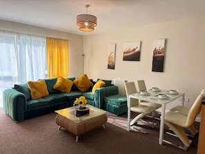 Una sala de estar moderna con un sofá verde, cojines amarillos, una mesa de centro de cristal y una zona de comedor con una mesa y sillas blancas.