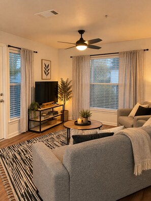 A living room with a sofa, a coffee table, a television, and a ceiling fan. - Between the Hedges | Cozy Poolside Escape (Athens)