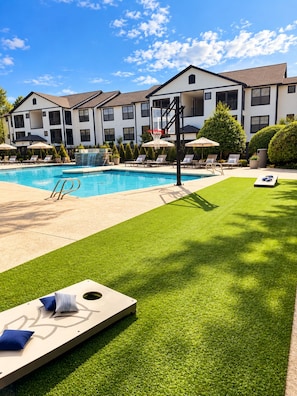 A swimming pool with a basketball hoop, surrounded by lounge chairs and umbrellas, with a building in the background. - Between the Hedges | Cozy Poolside Escape (Athens)