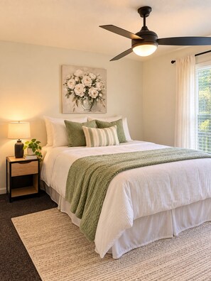 A bedroom with a bed, a nightstand, a lamp, a ceiling fan, a window with curtains, and a floral painting on the wall. - The Varsity Suite w/ Community Gym & Pool! (Athens)
