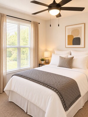 A bedroom with a bed, a ceiling fan, a window with curtains, a bedside table with a lamp, and a framed picture on the wall. - The Athens Playbook w/ Pool & Shuttle! (Athens)