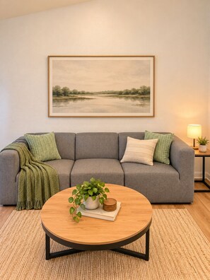 Ein Wohnzimmer mit einem grauen Sofa, einem runden Couchtisch mit einer Pflanze und einem gerahmten Landschaftsbild an der Wand.