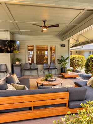 A covered outdoor seating area with a ceiling fan, a TV mounted on the wall, and a view of the house through large windows. - The Saturday Social w/ Shuttle and Amenities! (Athens)
