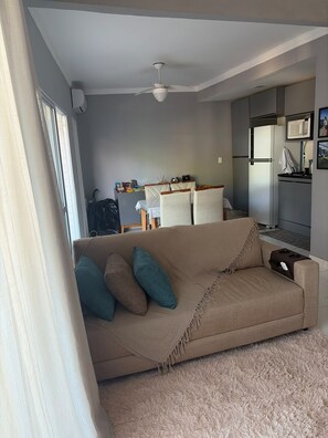 Una sala de estar con un sofá color beige, un ventilador de techo y una zona de cocina con microondas y refrigerador.