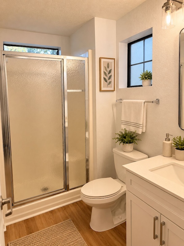 Een badkamer met een douchecabine van glas, een toilet, een wastafel met spiegel en een raam met een plant.