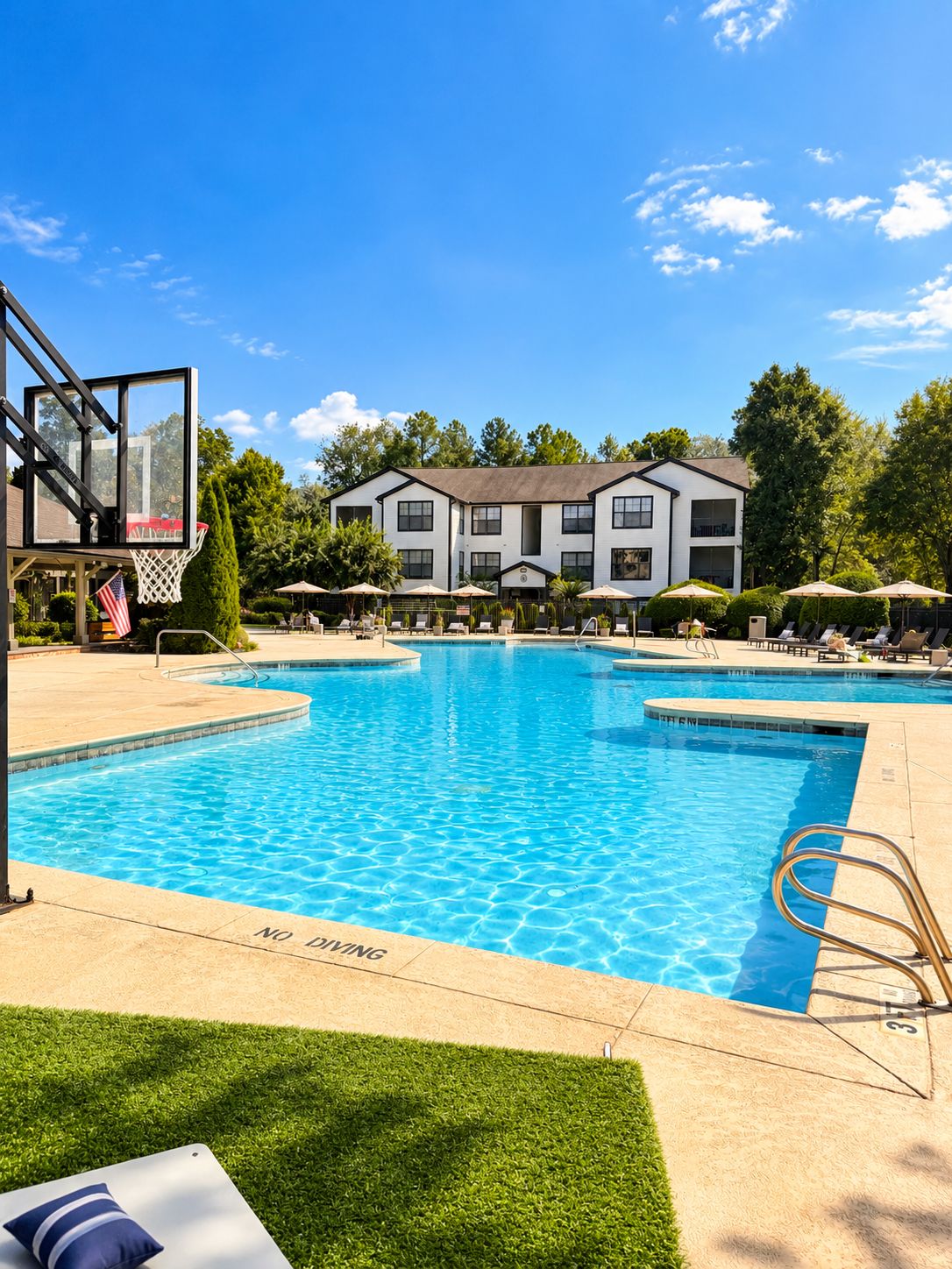 Ein Schwimmbecken mit einem Basketballkorb, Liegestühlen und einem Gebäude im Hintergrund.