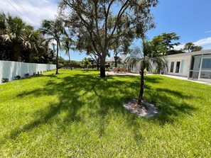 A well-maintained lawn with a patio area, a large tree, and a building with a modern design. - New Vacation Pool Home, 3 BD, Minutes from Beach! (Naples)