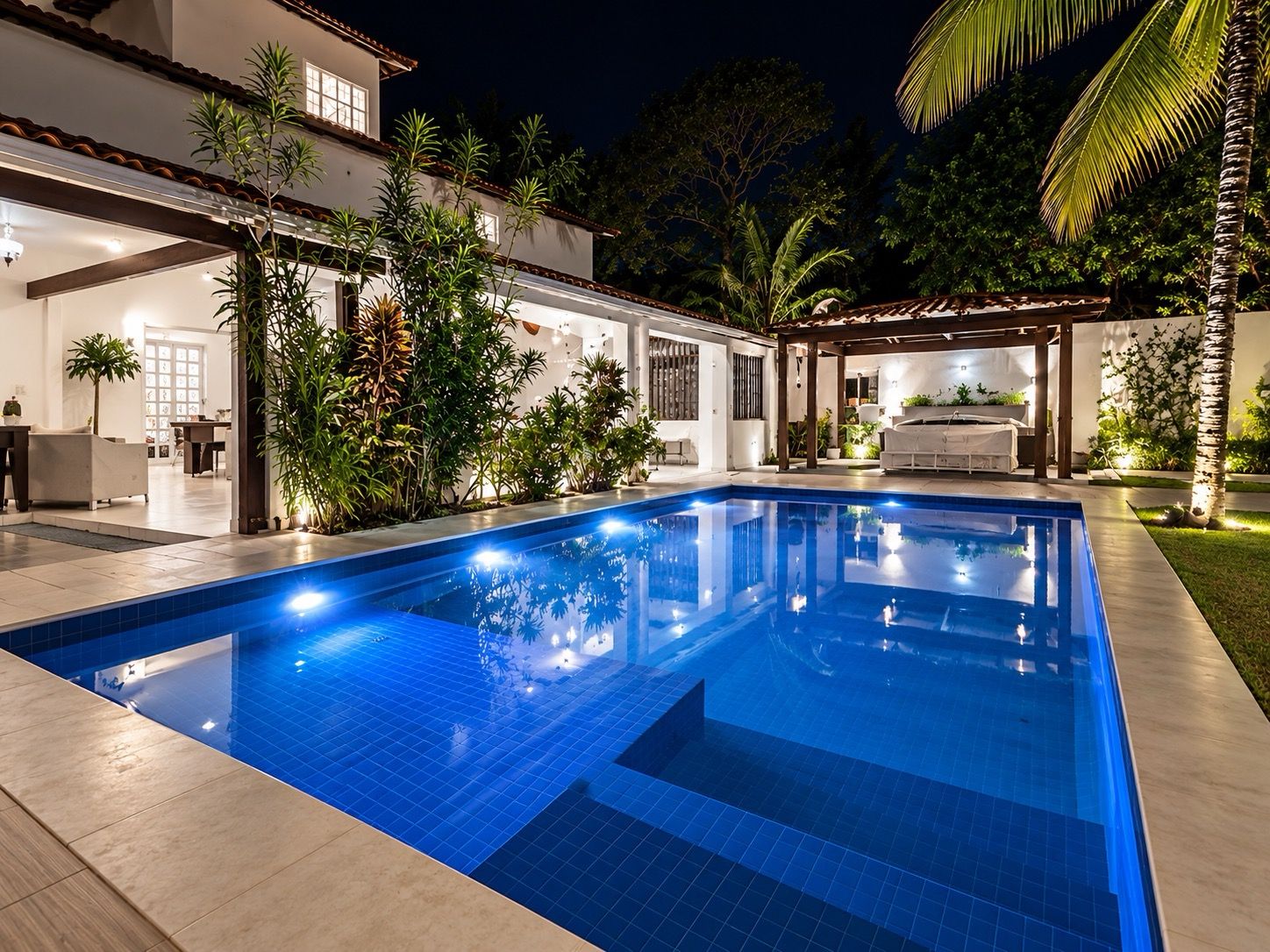 A swimming pool with a tiled deck, surrounded by a house with a covered patio and various plants.