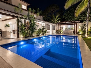 A swimming pool with a tiled deck, surrounded by a house with a covered patio and various plants. - New Listing (Arraial d'Ajuda)