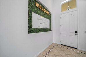 A hallway with a welcome sign, a white door, and a tiled floor. - All-Inclusive Retreat: Pool | HotTub | Golf | Games (Indio)