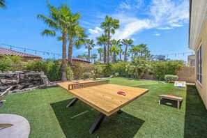 Una mesa de ping pong en un patio con césped artificial, palmeras y un muro de piedra.