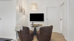 Ein modernes Wohnzimmer mit Flachbildfernseher an der Wand, zwei braunen Ledersesseln, einem runden Couchtisch und einer dekorativen Leuchte.