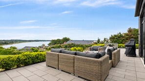 Een terras met rieten meubels, een glazen tafel en uitzicht op een rivier en gebouwen.