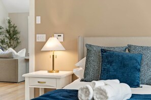 A neatly made bed with blue and white pillows, a bedside lamp, and a white nightstand with a drawer. - The Lodge at Woodland Acres (Taylors)