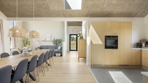 A modern kitchen with built-in appliances, a dining area with a wooden table and chairs, and a view through an open door to a garden. - VillaVilla 1023 - Hasmark Strand, Fyn og øer (Otterup)