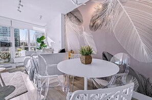 Uma sala de estar moderna com uma mesa redonda branca, cadeiras transparentes e uma grande janela com vista para a cidade.