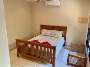 Ein Schlafzimmer mit einem hölzernen Bett, einer roten Tagesdecke, einem kleinen Holztisch und einer Klimaanlage an der Wand.