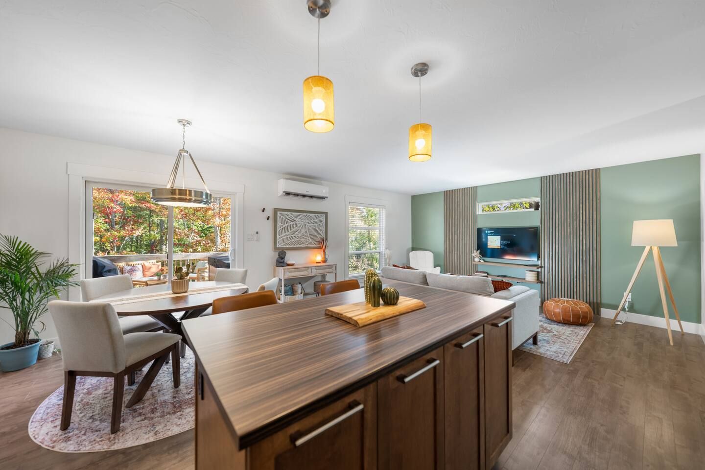 A modern kitchen with a central island, dining area with chairs, a large window with a view, and a television in the living area.