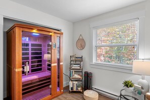 A room with a sauna, a bookshelf, a window with blinds, and a small table with a plant. - Serenity City Retreat•Shine & Rise Wellness (Moncton)