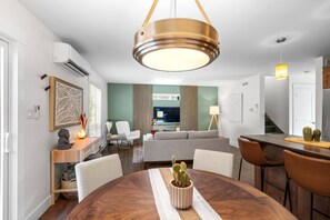 A modern living room with a dining area, a flat-screen TV, and a staircase leading to the kitchen. - Serenity City Retreat•Shine & Rise Wellness (Moncton)