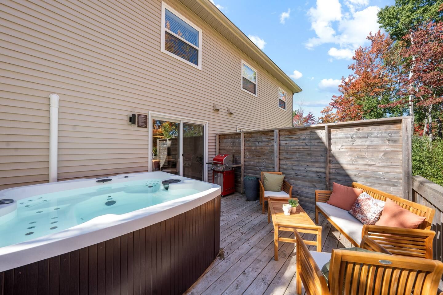 A hot tub on a wooden deck with outdoor furniture, a barbecue grill, and a house with siding and windows.