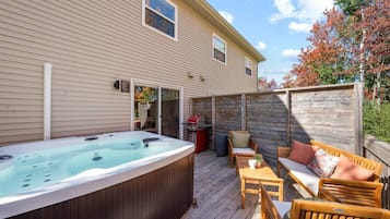 A hot tub on a wooden deck with outdoor furniture, a barbecue grill, and a house with siding and windows.