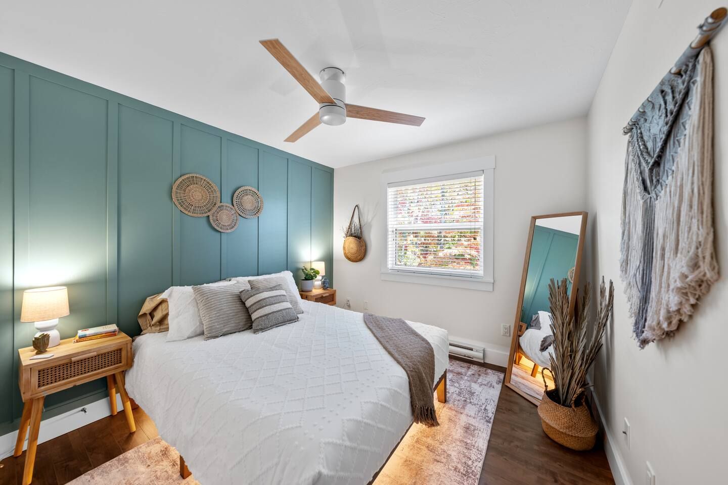 A bedroom with a bed, a ceiling fan, a window with blinds, a bedside table with a lamp, and a wall-mounted mirror.