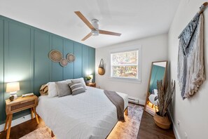 A bedroom with a bed, a ceiling fan, a window with blinds, a bedside table with a lamp, and a wall-mounted mirror. - Serenity City Retreat•Shine & Rise Wellness (Moncton)