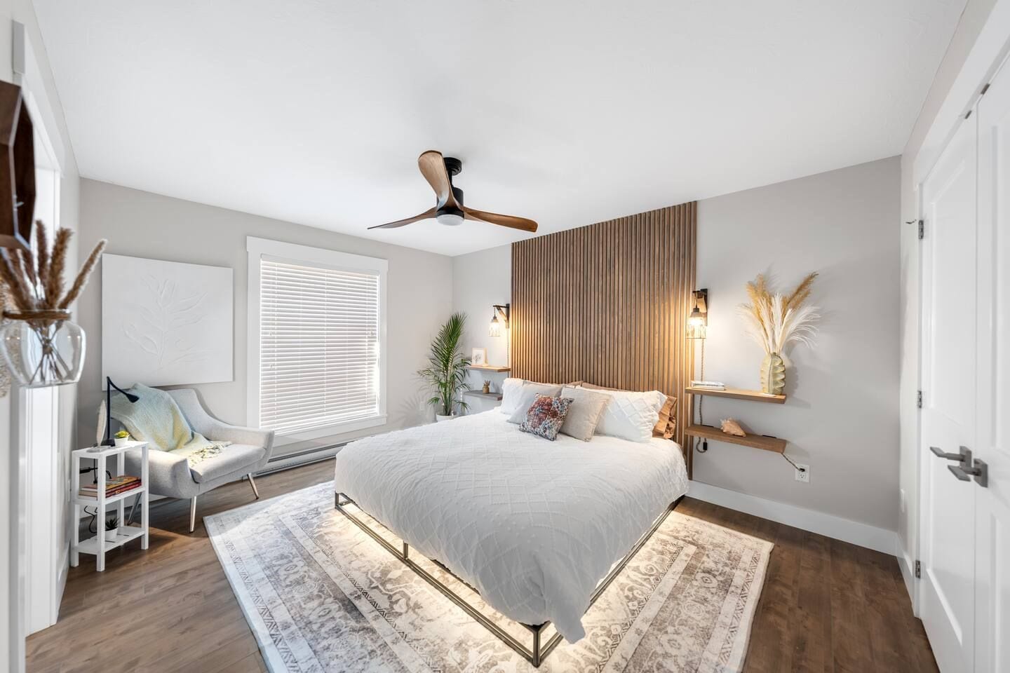 A modern bedroom with a large bed, a ceiling fan, a bedside table with a lamp, a window with blinds, and a decorative wall panel.
