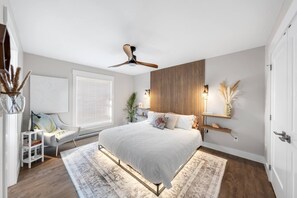 A modern bedroom with a large bed, a ceiling fan, a bedside table with a lamp, a window with blinds, and a decorative wall panel. - Serenity City Retreat•Shine & Rise Wellness (Moncton)