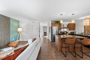 A modern kitchen with a central island, bar stools, and a dining area with a sofa. - Serenity City Retreat•Shine & Rise Wellness (Moncton)