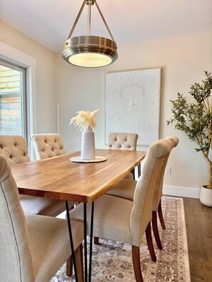 A dining area with a wooden table, cushioned chairs, a hanging light fixture, and a wall-mounted artwork. - Radiance City Retreat•Shine & Rise Wellness (Moncton)