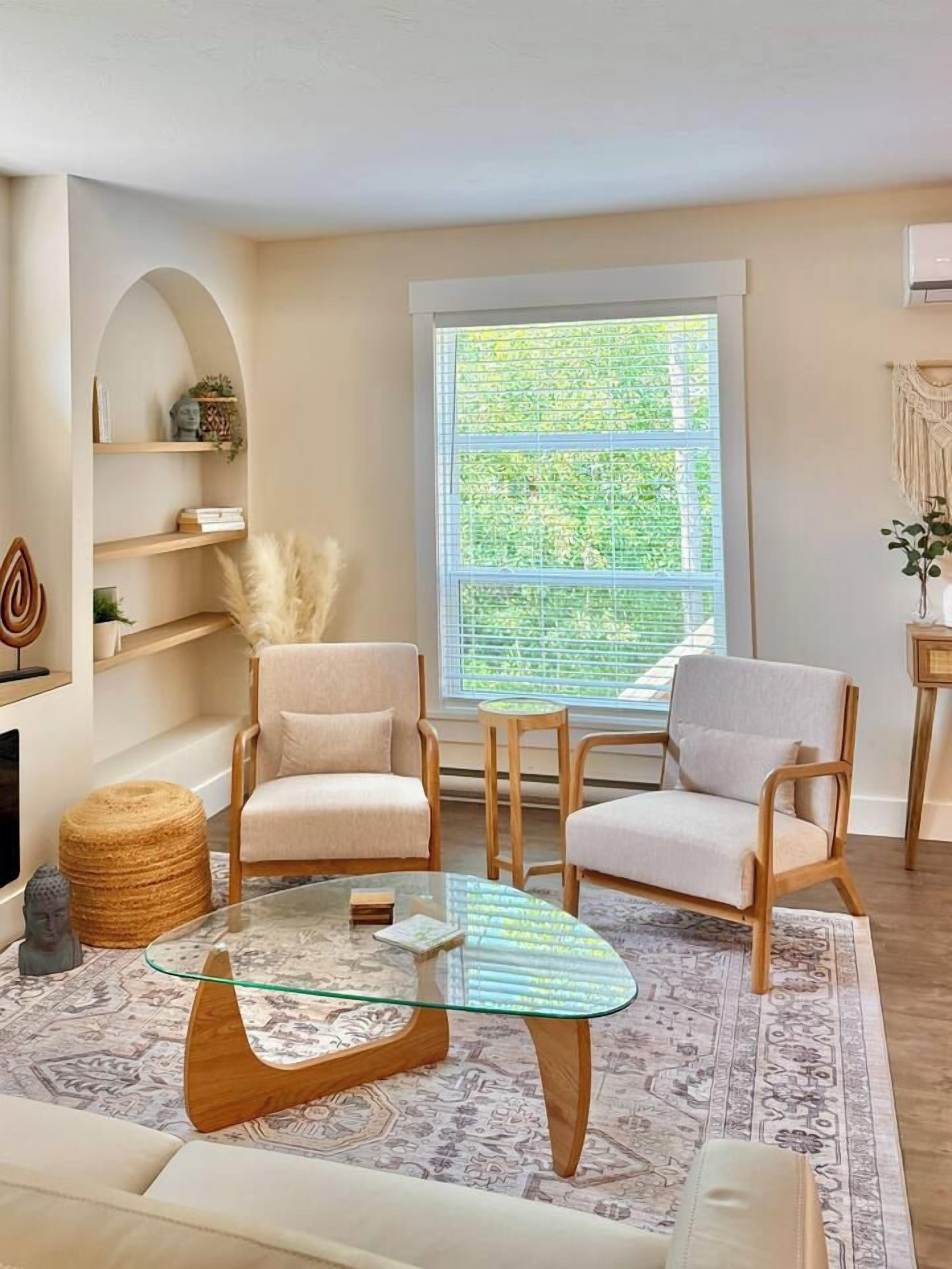 A living room with two armchairs, a glass coffee table, a patterned rug, and a large window with a view of greenery.