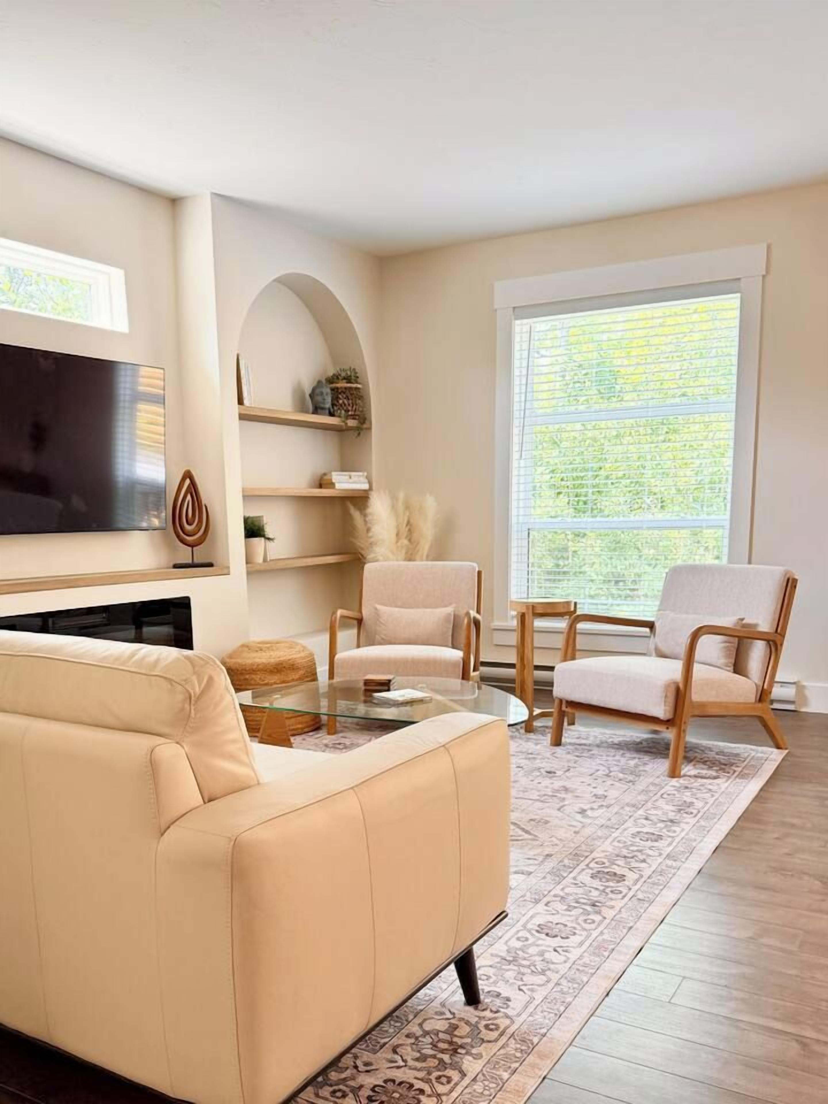 A living room with a beige sofa, two armchairs, a coffee table, a flat-screen TV, and a large window with blinds.