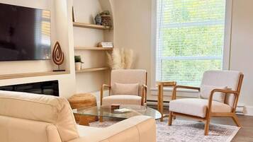 A living room with a beige sofa, two armchairs, a coffee table, a flat-screen TV, and a large window with blinds.