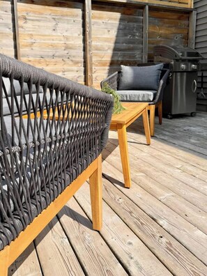A wooden deck with a wicker chair and a table, a grill, and a wooden wall in the background. - Radiance City Retreat•Shine & Rise Wellness (Moncton)