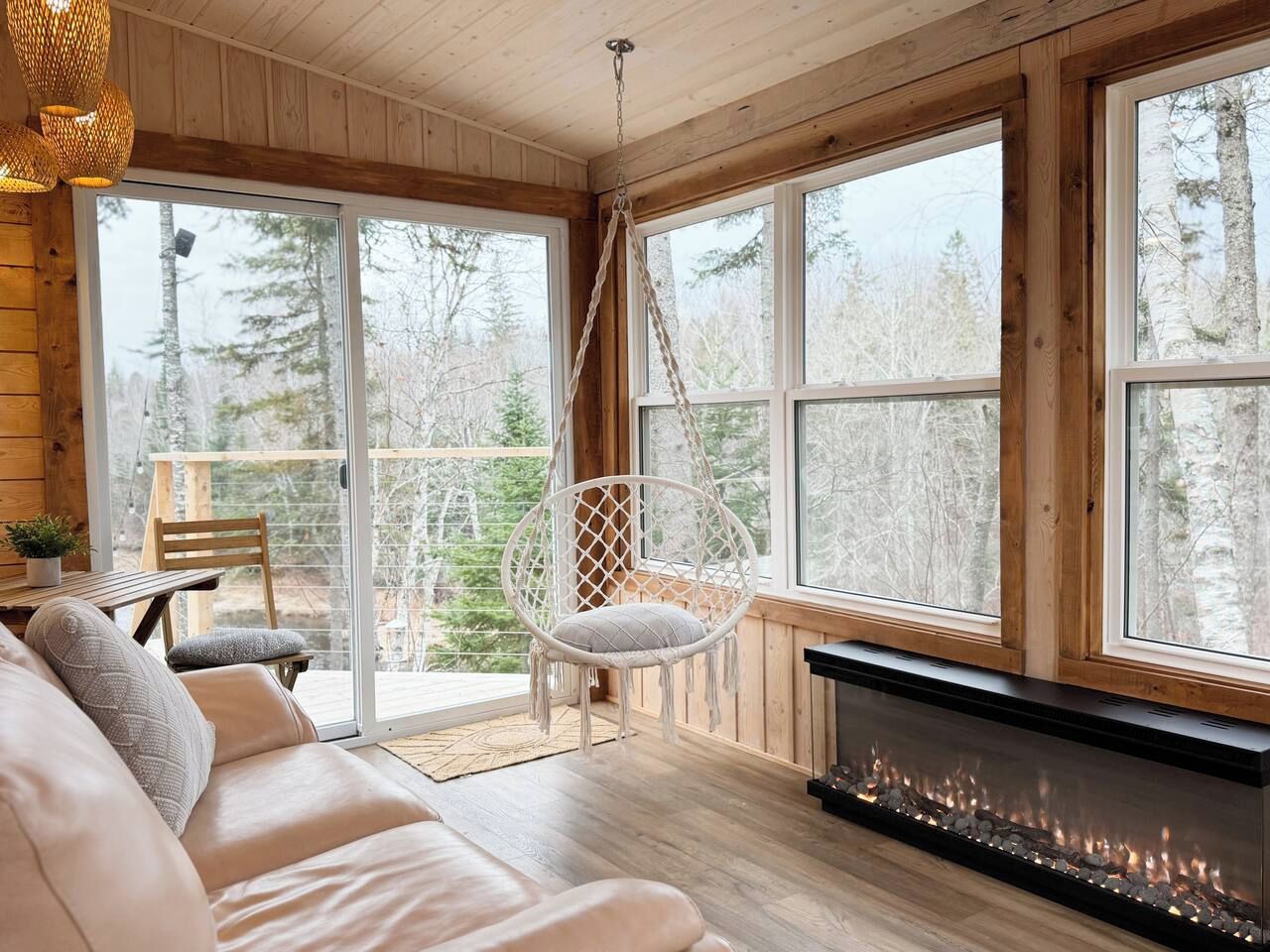 Interior acollidor d'una cabana amb un sofà, una cadira penjant, una llar de foc i grans finestres que donen vista a arbres.