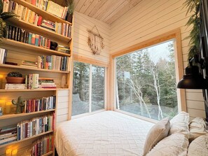 A cozy room with a large window, a bookshelf filled with books, and a bed with pillows.