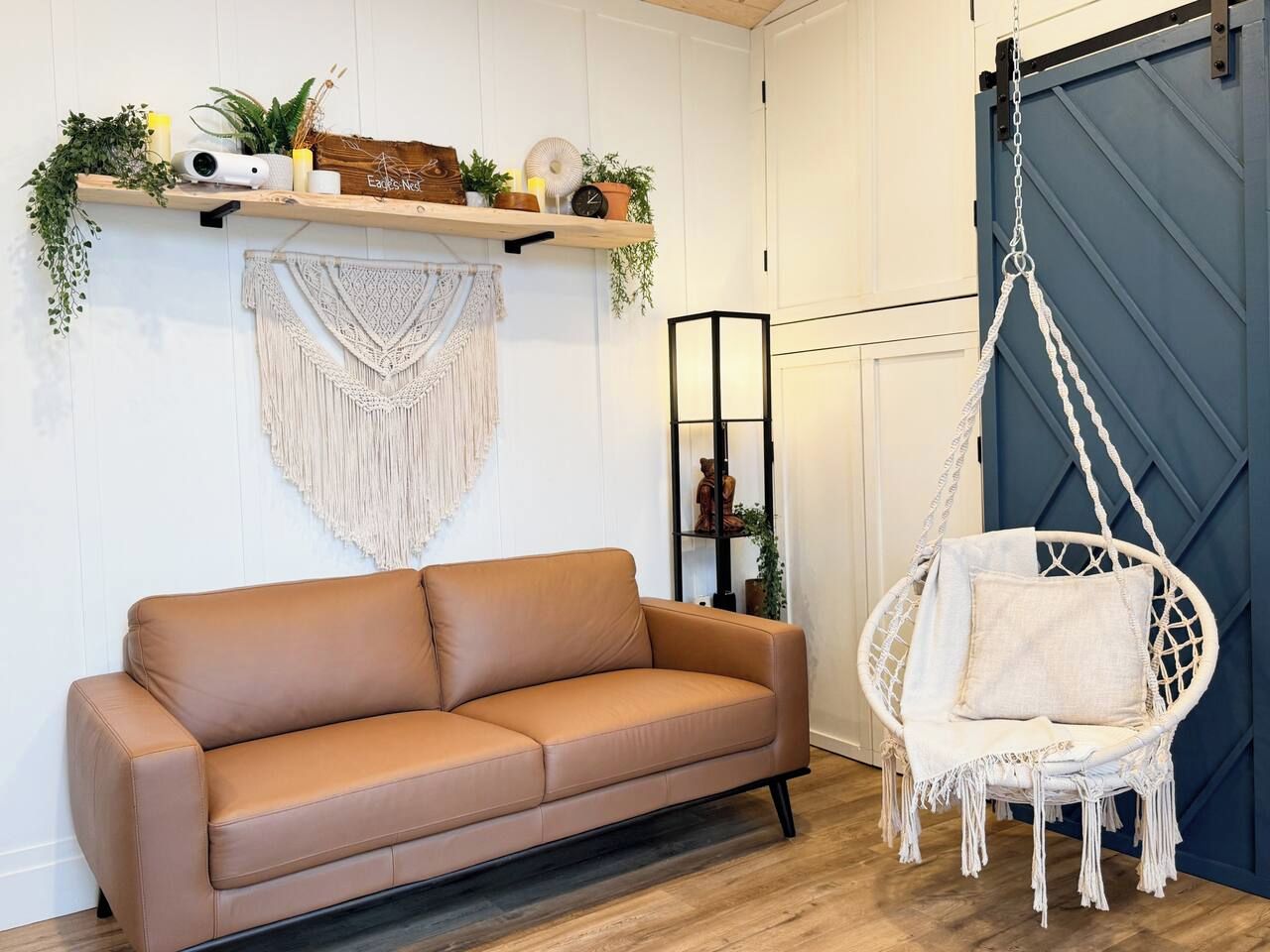 Un sofá de cuero color bronceado, un tapiz de macramé, una silla colgante y un estante con plantas y objetos decorativos.