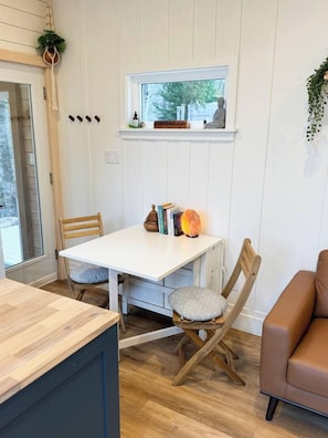 A room with a white table, two chairs, a brown sofa, a window with a view, and a small shelf with items. - Eagle’s Nest Nature Retreat•Shine & Rise Wellness (Saint Mary Parish)