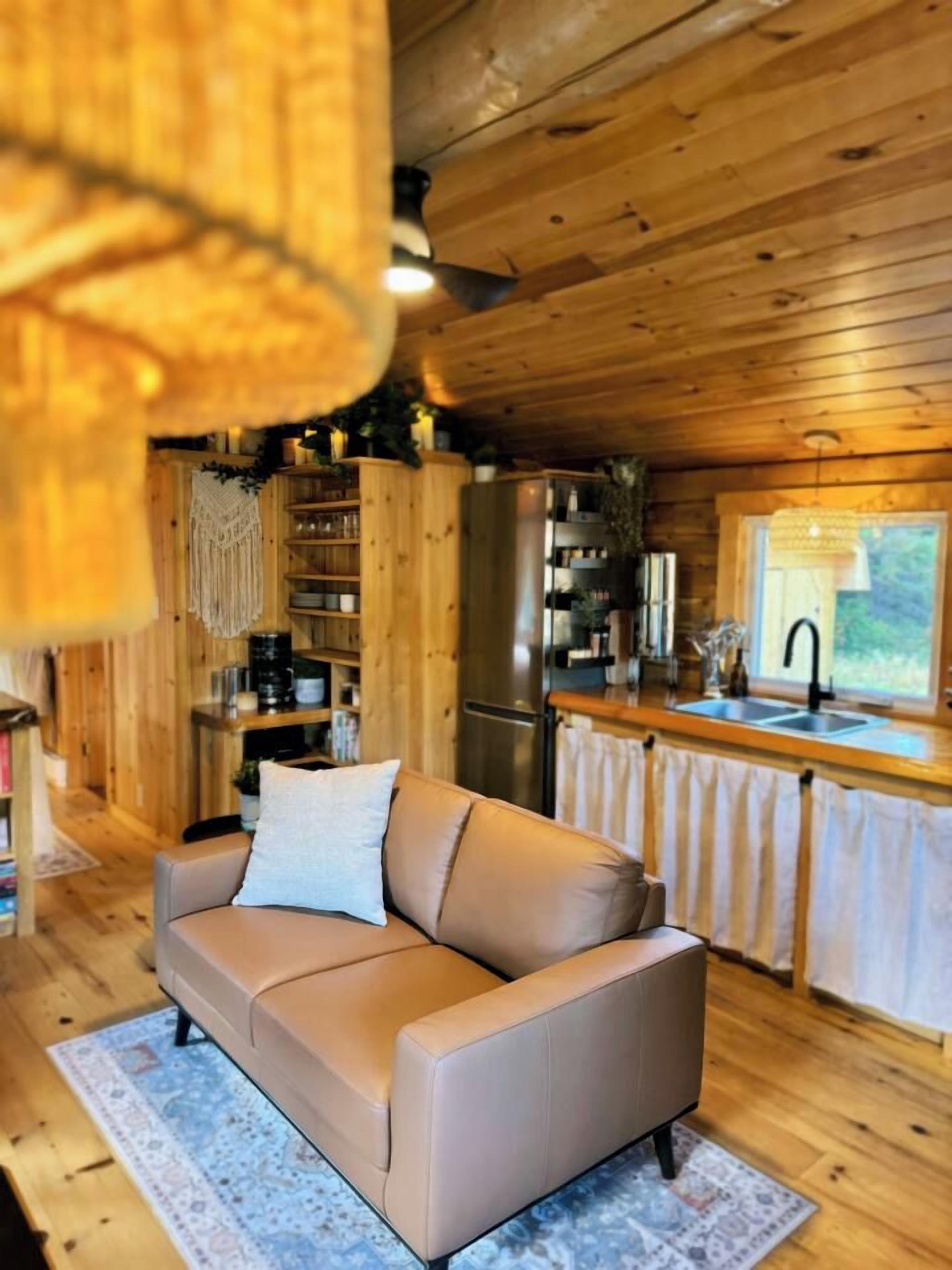 A cozy cabin interior with a sofa, wooden walls, a kitchen area, and a window with a view.