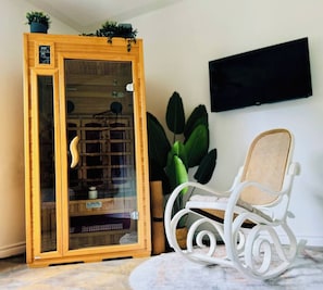A room with a wooden cabinet, a white rocking chair, a large green plant, and a flat-screen TV mounted on the wall. - Harmony Nature Retreat•Shine & Rise Wellness (Saint Mary Parish)