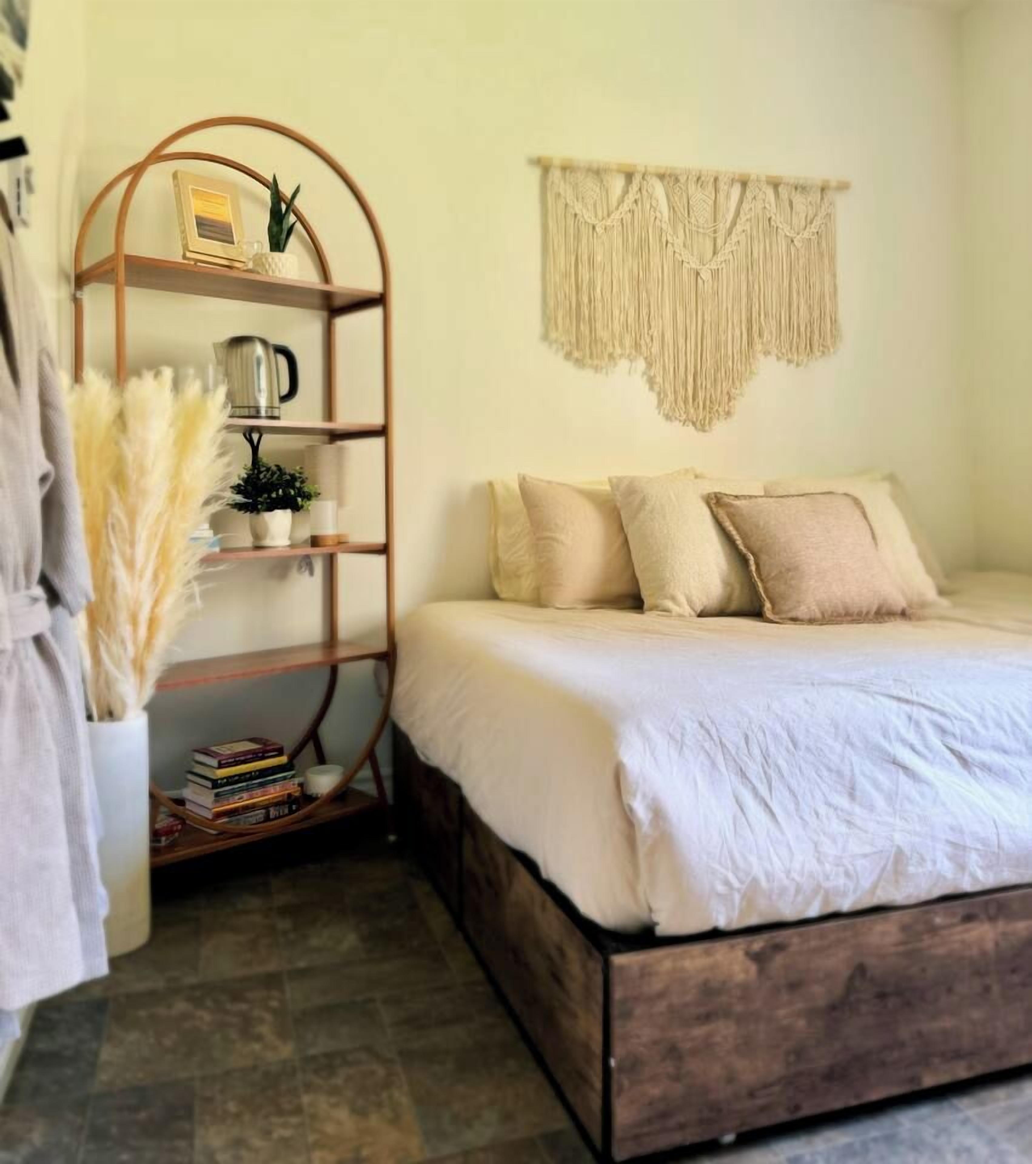 A bedroom with a bed, a wooden shelf with books, a macrame wall hanging, and a potted plant.